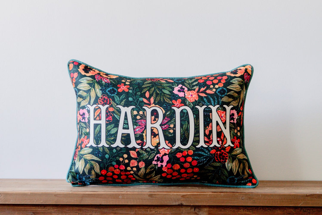 Floral pillow with embroidered name 'HARDIN' on a wooden surface against a white wall.